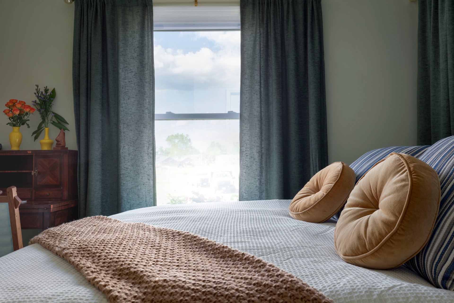 Bed foot with knit throw, velvet bolsters, and a view through the window to a cloudy sky